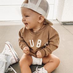 a toddler sitting on the ground wearing a brown shirt and hat with white letters