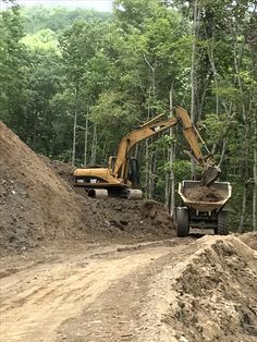 two construction trucks are parked on the side of a dirt road with trees in the background