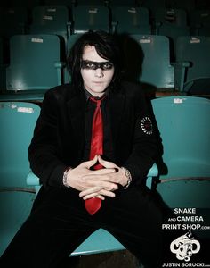 a man in a black suit and red tie sitting on a blue chair with his hands crossed