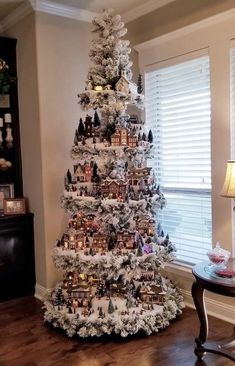 a white christmas tree with houses and trees on it in a living room next to a window