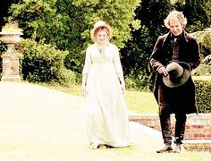 a man and woman dressed in period clothing walking down a path with trees in the background