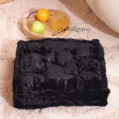 a black square pillow sitting on top of a white rug next to fruit and a bowl