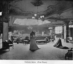 an old black and white photo of a woman in a formal dress looking at furniture