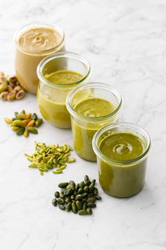 three jars filled with different types of green smoothies and pistachio seeds on a marble surface