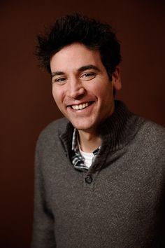 a smiling man with black hair wearing a sweater and looking at the camera while standing in front of a brown background