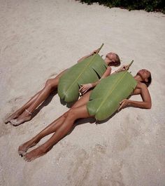 two women laying in the sand on their stomachs with large green leaves attached to them