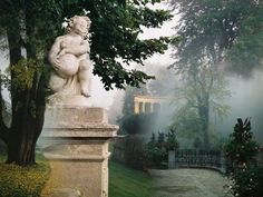 a statue sitting on top of a stone pillar next to a lush green park filled with trees