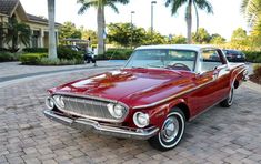 an old red car is parked in front of some palm trees and bushes on the sidewalk