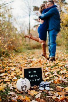 an engaged couple kissing in the woods surrounded by fallen leaves and pumpkins with a sign that says, one my gourd it's a boy