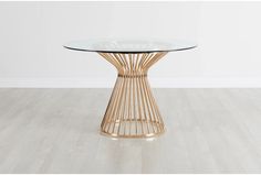 a round glass table with gold metal base on a wooden floor in front of a white wall