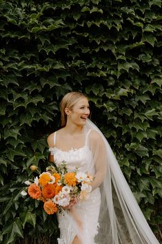 a woman standing in front of a bush holding a bouquet of flowers and a veil