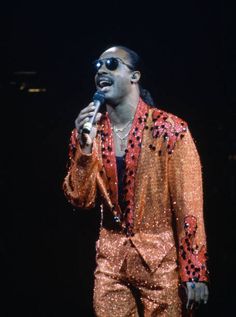 a man in an orange sequin suit holding a microphone and singing into a mic