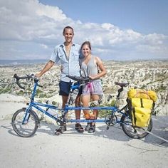 two people standing next to a blue bike on top of a rock covered hillside with trees in the background