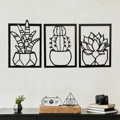 three black metal wall art pieces on a white wall next to a table with books and plants