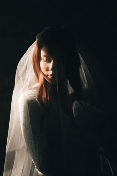 a woman with red hair wearing a white veil and looking down at her dress while standing in the dark