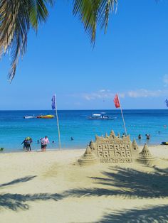 a sand castle on the beach with people in the water and sailboats in the background