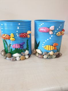 two blue tin cans with fish painted on them sitting next to rocks and pebbles in the water