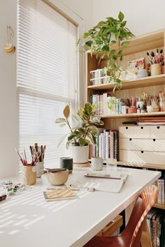 a white desk topped with lots of books next to a window filled with plants and office supplies