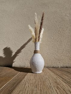 a white vase sitting on top of a wooden floor next to a wall with long grass sticking out of it