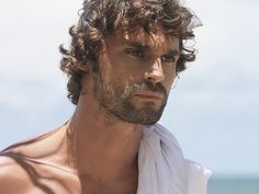 a man with curly hair and no shirt on standing by the ocean looking off into the distance