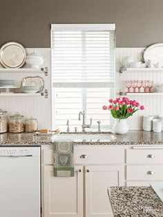 a kitchen with white cabinets and marble counter tops, pink flowers in a vase on the window sill