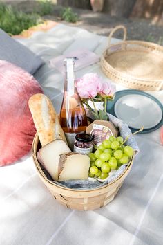 a picnic basket with wine, cheese and bread on a blanket outside in the sun