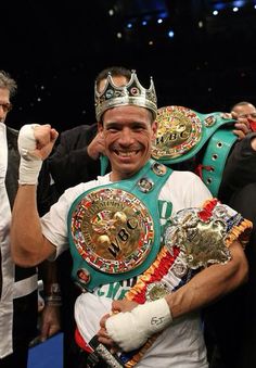 a man wearing a crown and holding two boxing belts in front of his face with other people around him