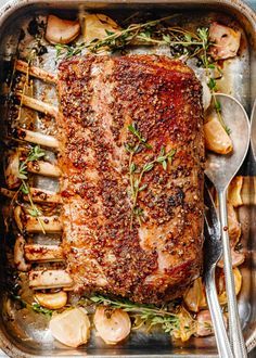 a roasting pan filled with meat, potatoes and herbs next to spoons on a table