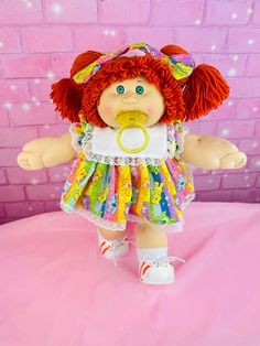 a small doll with red hair wearing a dress and pacifier in front of a pink brick wall