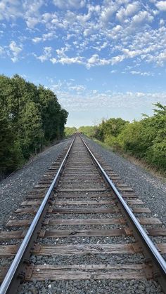 an empty train track surrounded by trees and bushes under a blue sky with wispy clouds