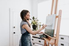 a woman is painting on an easel in her living room while holding a paintbrush