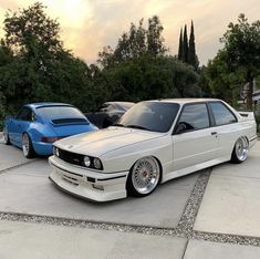 two cars parked next to each other in a parking lot with trees and bushes behind them