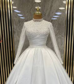 a white wedding dress with long sleeves and beading on the neckline is displayed in front of a marble wall