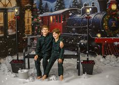 two young boys sitting on a bench in front of a christmas train backdrop with snow