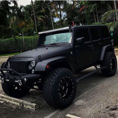 a black jeep parked in a parking lot next to some palm trees and bushes on the side of the road