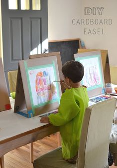 a little boy sitting at a table with some paintings on it and an easel in front of him