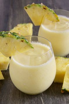 two glasses filled with pineapple drink sitting on top of a wooden table next to sliced pineapples