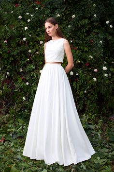 a woman standing in front of flowers wearing a white dress with cropped top and long skirt