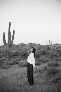 a woman standing in the middle of a desert with cacti and cactus behind her