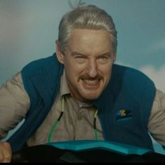 a man with grey hair wearing a blue vest and smiling at the camera while sitting down