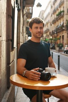 a man sitting at a table with a camera in front of him and holding a coffee cup