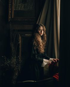 a woman with long hair sitting on a chair in front of a window wearing a black dress