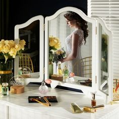 a woman standing in front of a white vanity with flowers and makeup products on it