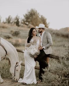 a bride and groom sitting on a fence next to a white horse in the field