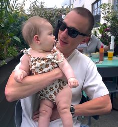 a man holding a baby in his arms while sitting at an outdoor table with plants