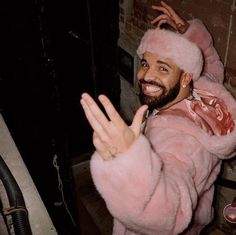 a man wearing a pink fur coat and holding his hand up in the air while standing next to a brick wall