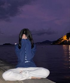 a woman sitting on the edge of a body of water with her head in her hands