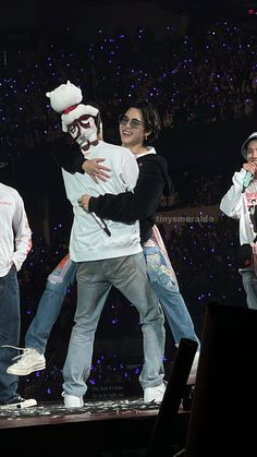 two men are hugging on stage with other people in the background and one is wearing a clown mask