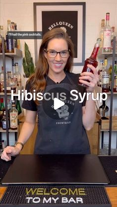 a woman holding up a bottle in front of a welcome to my bar sign that says hibiscus syrup