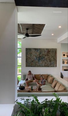 two people sitting on a couch in a living room with plants and potted plants